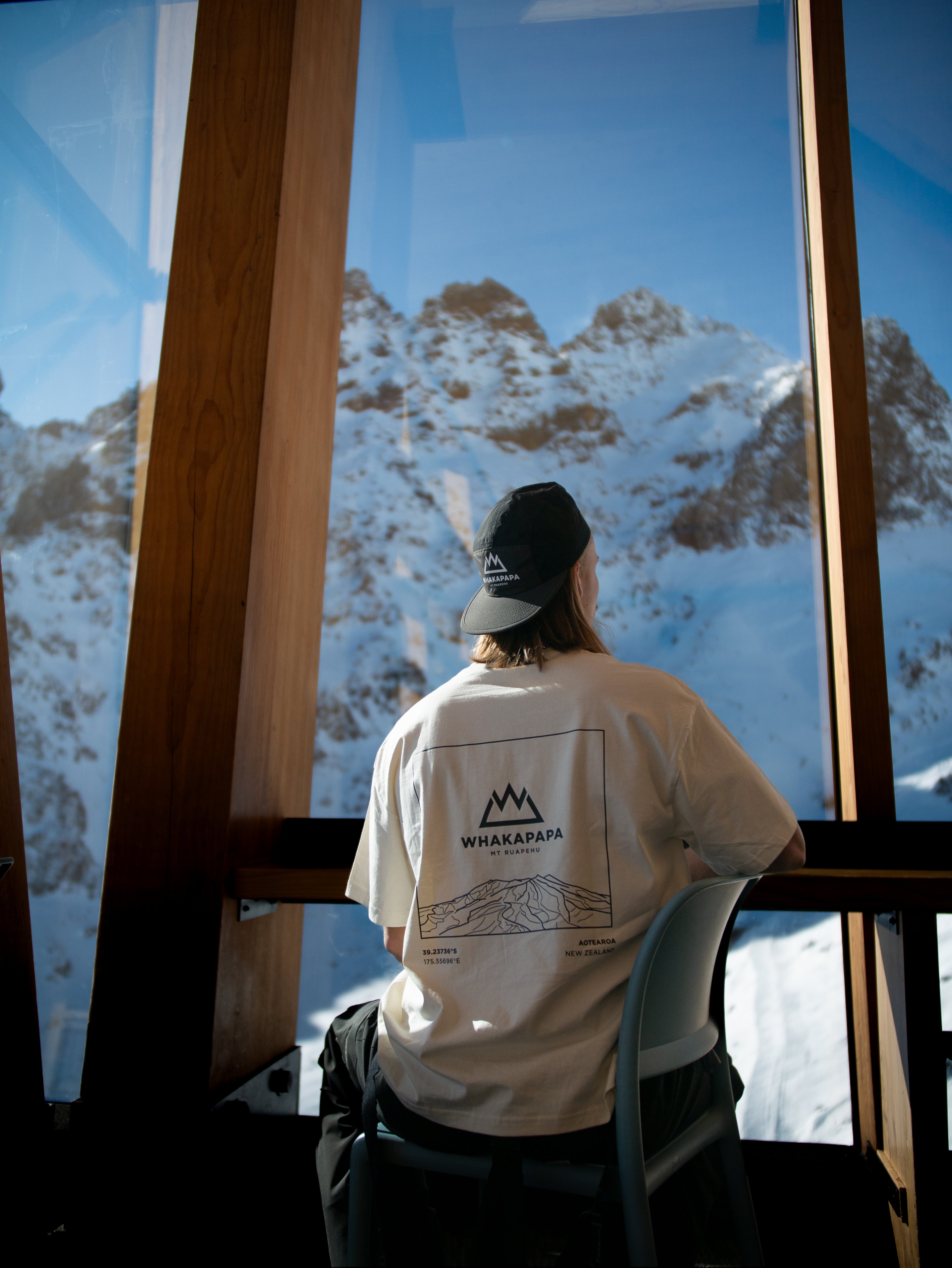 Person wearing a white t-shirt with a mountain logo, sitting inside a building looking out at snow-covered mountains.